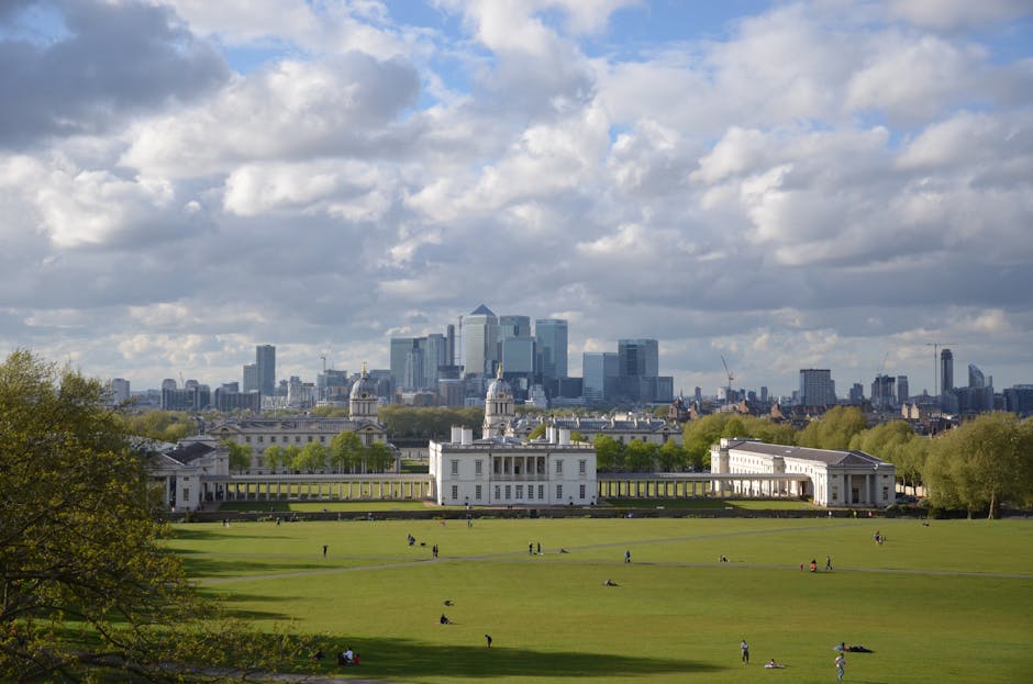 A wide panoramic view of Greenwich Park with its expansive green lawns dotted with people walking, sitting, and relaxing, bordered by rows of trees. In the foreground, there is a large white historic building with classical architectural features, including columns and a symmetrical façade, situated within the park. Beyond the park, the modern skyline of London, featuring numerous high-rise office buildings and skyscrapers, rises under a partly cloudy sky with patches of blue. The scene captures the contrast between the open green space and urban architecture, illustrating an area where local house removals and furniture transport services, such as those provided by Removal Van Greenwich, often operate to facilitate home relocation and logistics for residents and venues attending events in the city center.
