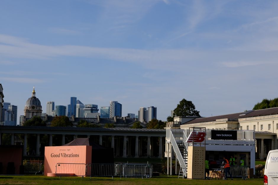 The image depicts an outdoor scene with a clear blue sky and a city skyline featuring modern tall buildings and a domed historic structure in the background. In the foreground, there is a temporary outdoor setup with a pink sign reading 'Good Vibrations' placed on the grass, surrounded by temporary metal barriers. To the right, a small structure with stairs leading up to a second level displays 'New Balance' branding, appearing to be a mobile stage or event booth. Several people wearing high-visibility vests are visible near the structure, engaged in activities that suggest loading or organizing equipment, possibly related to an event or relocated setup. Adjacent to the event area, the surroundings include a low, classical-style building with columns and a green landscape, indicating a park or open space within or near Greenwich. This scene captures the logistics involved in setting up or relocating event equipment as part of a house or venue move service provided by Removal Van Greenwich, aligned with their specialist removals and moving logistics services.