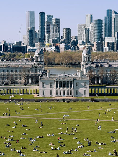 A wide panoramic view of Greenwich Park with its expansive green lawns dotted with people walking, sitting, and relaxing, bordered by rows of trees. In the foreground, there is a large white historic building with classical architectural features, including columns and a symmetrical façade, situated within the park. Beyond the park, the modern skyline of London, featuring numerous high-rise office buildings and skyscrapers, rises under a partly cloudy sky with patches of blue. The scene captures the contrast between the open green space and urban architecture, illustrating an area where local house removals and furniture transport services, such as those provided by Removal Van Greenwich, often operate to facilitate home relocation and logistics for residents and venues attending events in the city center.