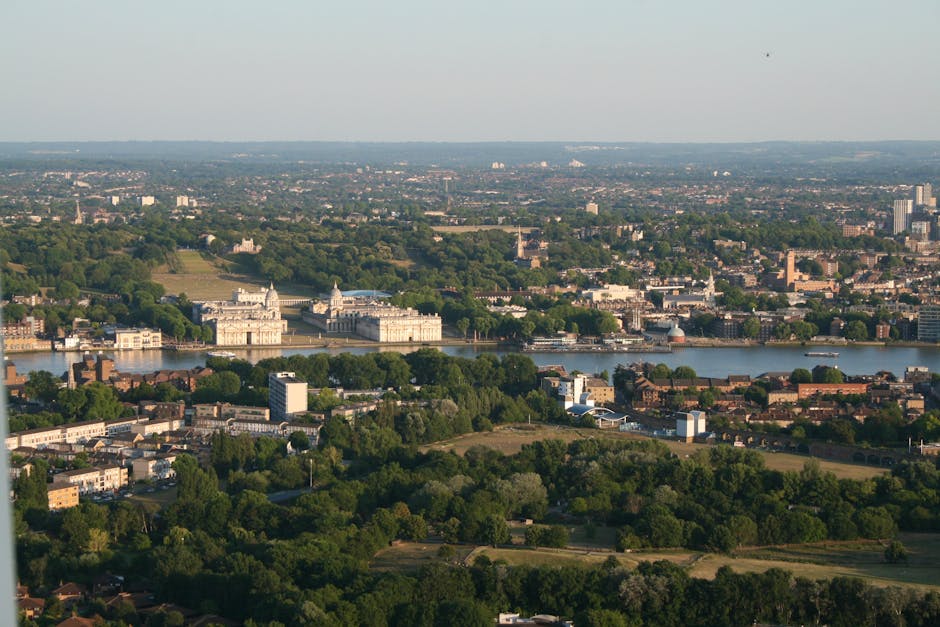 A wide outdoor view of Greenwich Park in London during daytime, with a large green lawn in the foreground where numerous people are sitting and lying on the grass. In the middle ground, the classical 
