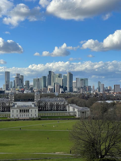 A panoramic view of central London showing a large, open green park in the foreground with a few people walking across the grass. Behind the park, there is a historic white building with classical architecture, featuring columns and a symmetrical facade. Further back, a modern city skyline dominates the scene, with numerous high-rise office buildings and residential towers made of glass and steel under a partly cloudy blue sky. The image captures the contrast between the expansive green space and the dense urban development, illustrating a typical setting where house removals and furniture transport services by Removal Van Greenwich might occur during a home relocation or moving process in Greenwich.