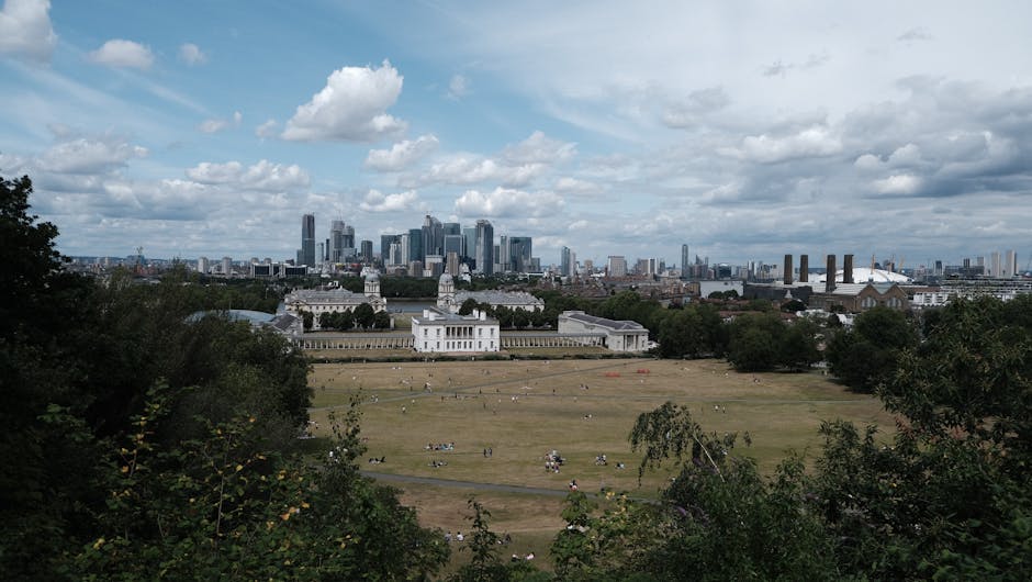 A wide view of a park in Greenwich, showing a large open grassy area with several groups of people relaxing and picnicking. In the middle distance, a white historic building with classical architecture is visible, surrounded by trees and pathways. Beyond the park, the modern skyline of tall office buildings and skyscrapers rises against a partly cloudy sky with patches of blue. To the right, some industrial structures and factory chimneys are seen in the background. The scene is captured from an elevated vantage point, with some foliage framing the foreground. This outdoor setting relates naturally to home relocation and moving logistics, as it highlights the environment where furniture or boxes might be loaded into a removals vehicle, such as those operated by Removal Van Greenwich, during packing and moving processes in Greenwich.