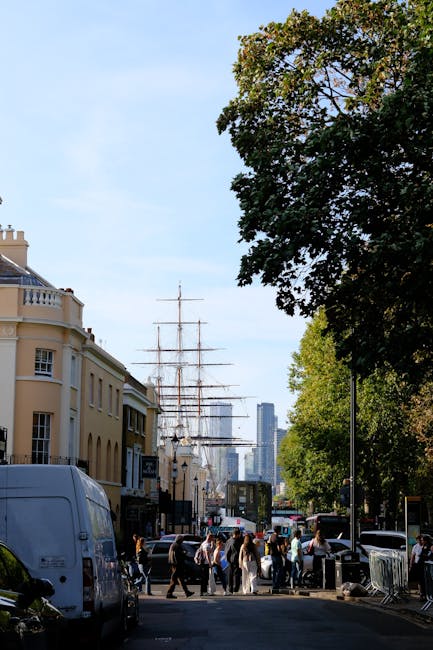 A street scene in Greenwich with a tall sailing ship docked nearby, identifiable by its masts and rigging, visible behind modern skyscrapers. On the street, a group of people are walking across the pavement, some carrying shopping bags, while others stand near parked cars and a white delivery van. To the right, mature trees with green foliage shade part of the sidewalk, and a black vehicle is parked along the curb. In the background, a large historic house with cream-coloured walls and decorative railings on its balcony is adjacent to the street. The scene is outdoors under a clear blue sky, capturing the lively environment of Greenwich, with visible details relevant to local moving and transport activities, such as the parked vehicles and pedestrians involved in the movement process. Removal Van Greenwich is familiar with this environment, providing home relocation and furniture transport services near Cutty Sark.