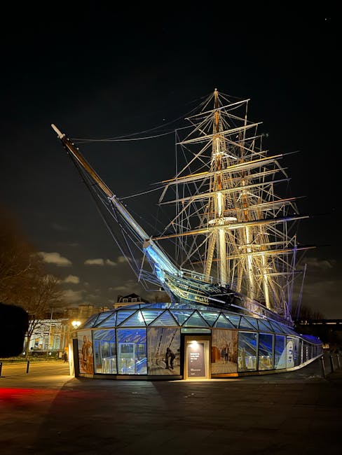 Night view of the Cutty Sark maritime museum in Greenwich, London, showing the illuminated historic sailing ship's wooden masts, rigging, and hull structure. The ship is positioned on a glass-paned exhibition space that surrounds its base, with the reflections of lights visible on the transparent surfaces. The background includes a dark cloudy sky with a few stars, and distant city lights. The setting highlights the ship's museum environment, which is located near the river and accessible for visitors. This image is relevant to Greenwich's historical attractions and the area's transportation and touring infrastructure, complementing content about house and furniture removals in Greenwich in proximity to local landmarks like the Cutty Sark.