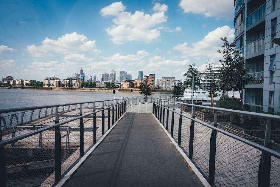 The image displays a metal walkway with safety railings extending over a body of water, leading towards the city skyline in the distance. On either side of the walkway, there are trees and modern resi