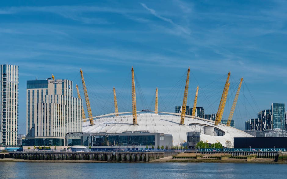 The image displays a metal walkway with safety railings extending over a body of water, leading towards the city skyline in the distance. On either side of the walkway, there are trees and modern resi