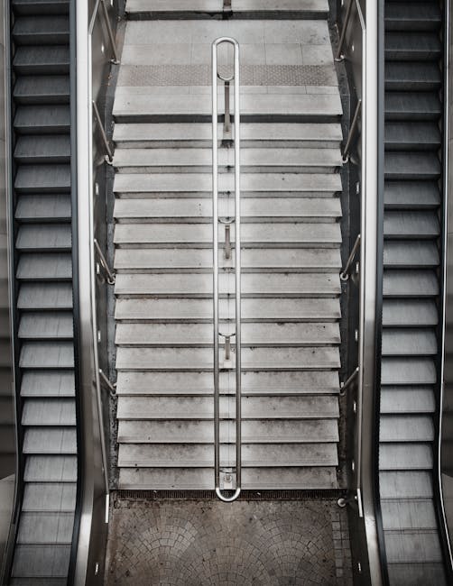 A straight indoor staircase with black steps and yellow safety edges, leading upward within a building structure. The staircase is flanked by yellow handrails on both sides, supported by metal balustr