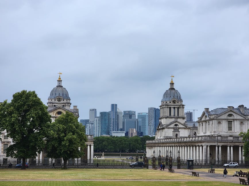 The image shows a spacious outdoor area in Greenwich with historic buildings featuring domed roofs and classical architectural elements in the foreground, with a large green tree partially obscuring parts of these structures. Behind the historic buildings, modern high-rise office skyscrapers rise into a cloudy sky, creating a stark contrast between old and new architecture in the city skyline. The open grassy area in the foreground appears to be a park or lawn, with a paved walkway and a few benches along the perimeter. In the scene, a removal van belonging to Removal Van Greenwich is parked on the pavement near the buildings, with visible loading equipment such as trolleys and packing boxes nearby, indicating a home relocation or furniture transport operation underway. The lighting is natural and diffuse due to the overcast sky, providing a clear, detailed view of the entire setting consistent with professional moving and packing activities.