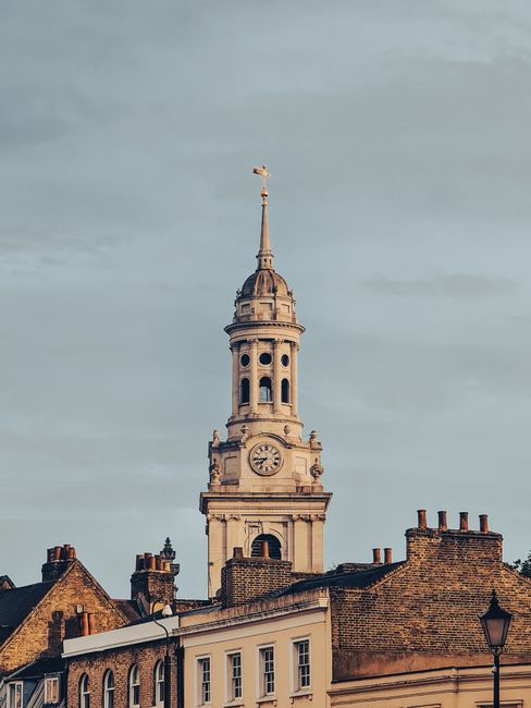 A tall historic clock tower with a pointed spire, situated among traditional brick buildings with chimneys, is visible against a cloudy sky during daytime. The clock on the tower shows the time, and the structure features ornate architectural details. In the foreground, the tops of residential buildings with tiled roofs and a street lamp are seen. This scene represents a typical urban setting in Greenwich, London, relevant to house removals and furniture transport services. The image captures the loading area outside a property, where a removal van may be parked nearby, ready for home relocation, with furniture and packing materials possibly being prepared for transport by Removal Van Greenwich, indicating a professional moving process involving loading and packing in preparation for a smooth move.