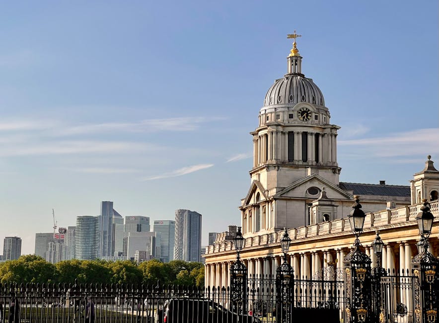 The image shows a spacious outdoor area in Greenwich with historic buildings featuring domed roofs and classical architectural elements in the foreground, with a large green tree partially obscuring parts of these structures. Behind the historic buildings, modern high-rise office skyscrapers rise into a cloudy sky, creating a stark contrast between old and new architecture in the city skyline. The open grassy area in the foreground appears to be a park or lawn, with a paved walkway and a few benches along the perimeter. In the scene, a removal van belonging to Removal Van Greenwich is parked on the pavement near the buildings, with visible loading equipment such as trolleys and packing boxes nearby, indicating a home relocation or furniture transport operation underway. The lighting is natural and diffuse due to the overcast sky, providing a clear, detailed view of the entire setting consistent with professional moving and packing activities.