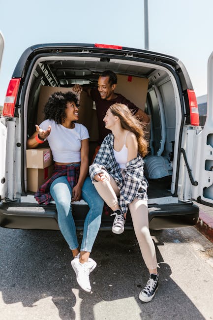 Two young women sit inside the open rear of a white moving van, engaged in conversation and smiling. One woman has curly black hair, is wearing a white T-shirt and denim jeans, and has a plaid shirt tied around her waist. The other woman has long, straight, reddish-brown hair, and is wearing a white tank top with a plaid shirt draped over her shoulders and light-colored tights. The interior of the van contains several cardboard boxes, some wrapped in plastic, and appears well-organized for home relocation. The van's interior features a metal frame with horizontal bars for securing items, and the exterior is visible with a red safety light on top. The scene takes place outdoors in bright daylight, with the open rear door revealing a paved surface and a curb. This image exemplifies furniture transport, packing, and loading processes carried out by a professional removals service, such as Removal Van Greenwich, during a house move.