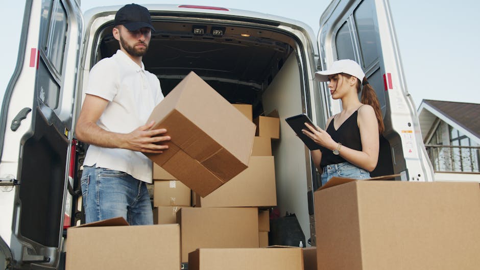 A young man wearing a black cap, white t-shirt, and blue jeans is inside a white moving van, handling a large cardboard box during the home relocation process. Several other cardboard boxes of various sizes are stacked around him within the van. On the right side, a young woman with a white cap, black sleeveless top, and denim shorts is standing outside the van, holding a portable device, possibly for inventory or instructions. The scene takes place outdoors on a paved street with a residential house visible in the background. The van's rear doors are open, revealing the interior filled with packed boxes and moving equipment such as straps and blankets, associated with professional removals. This setup reflects furniture transport and packing activities typical of house removals, as offered by Removal Van Greenwich, supporting efficient loading and moving logistics for clients.